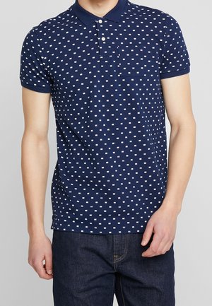 Man in een marineblauwe polo met korte mouwen met een wit klein vlaggenpatroon en donkerblauwe jeans.