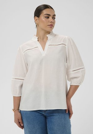 Moteris, tamsiais plaukais surištais atgal, vilki balta palaidine su nėrinių įsiuvais ir trijų ketvirčių pūstomis rankovėmis, derinančia su mėlynomis džinsomis.