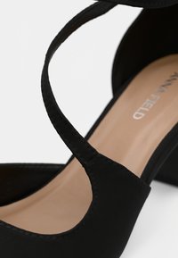 Črne visoke petke z prečnimi trakovi, gladek sintetični material, odprta stran in bež podplat s subtilnim znakom blagovne znamke.