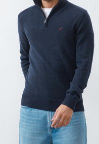 Man som bär en marinblå quarter-zip tröja med en liten röd logotyp på bröstet och ljusblå jeans, stående mot en vit vägg.