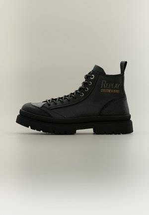 Botte montante en cuir noir avec semelle épaisse, lacets et texte "Replay Custom Service" sur le côté sur un fond uni.