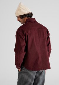 Hombre con gorro de punto color crema, chaqueta burdeos y pantalones gris a rayas, de pie con las manos en los bolsillos, mirando de lado contra un fondo blanco.