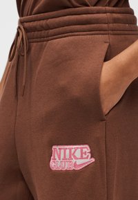 Καφέ φόρμα από απαλό ύφασμα, με ροζ κέντημα "NIKE CLUB" στο αριστερό τσέπη, με ρυθμιζόμενη μέση με κορδόνι.