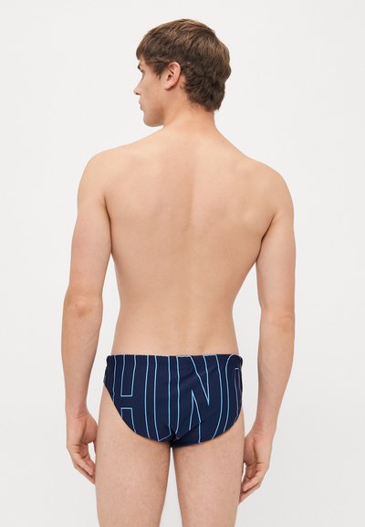 Junger Mann mit kurzem braunem Haar, der sich abwendet, trägt marineblaue Schwimmshorts mit großen, hellblau umrandeten Buchstaben auf der Rückseite.