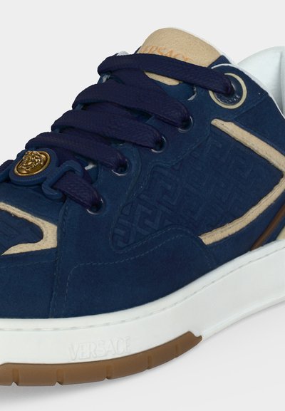 Κοντινό πλάνο ενός μπλε και μπεζ sneaker Versace με σκούρα μπλε κορδόνια, χρυσό έμβλημα Medusa, ανάγλυφο σχέδιο και λευκή σόλα με καφέ πέλμα.
