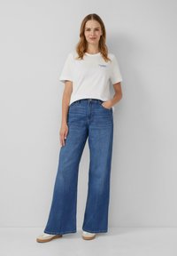 Witte t-shirt met blauwe tekst, gecombineerd met hooggerande, losse blauwe denim jeans. De jeans hebben een rechte pijp en subtiele vervaging.