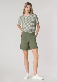 Pantaloni corti verdi con una vestibilità rilassata, abbinati a una t-shirt a righe bianche e olive con un piccolo emblema. Sneakers bianche completano l'outfit.