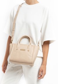 Beige leren handtas met korte handvatten, een afneembare schouderriem en een geprofileerde voorkant met kleine gouden accenten.