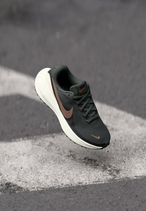 Czarno-brązowy but biegowy Nike w powietrzu nad asfaltem z białymi oznaczeniami ulicznymi, ukazujący cząsteczki brudu wokół podeszwy buta.