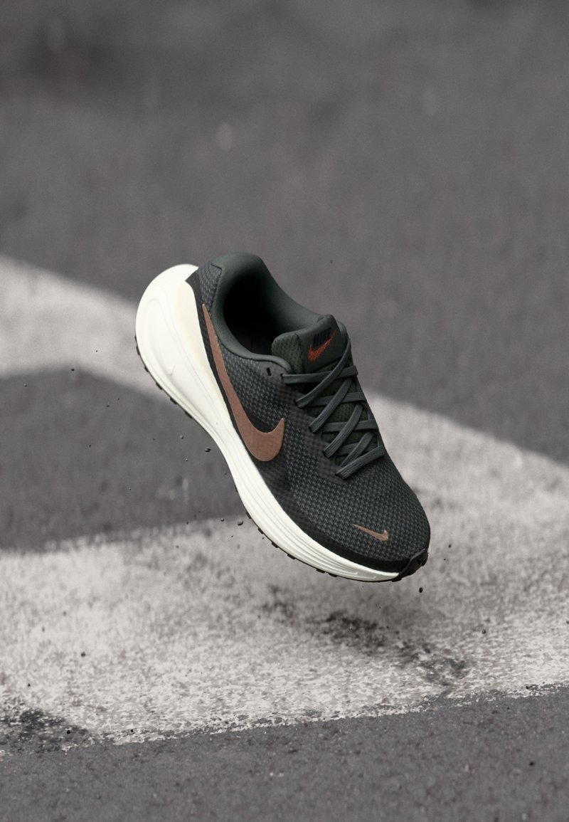 Chaussure de course Nike noire et marron en l'air au-dessus de l'asphalte avec des marquages blancs sur la route, montrant des particules de saleté autour de la semelle.