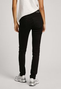 Person som bär slim svarta jeans, vita sneakers och en lös vit topp, står med ryggen mot betraktaren på en neutral bakgrund.