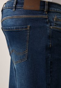 Siniset farkut, joissa on ruskea nahkatunnus, jossa lukee "MEN+", oranssit tikkausja ja standardin muotoiset takataskut.