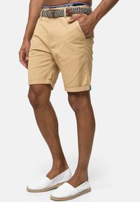 Shorts beiges avec des revers, poches avant et une ceinture tissée. Chaussures slip-on blanches avec une surface texturée et une semelle beige.