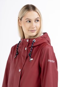 Giacca impermeabile rossa con cappuccio, caratterizzata da dettagli con logo bianco, cordini blu marina con accenti in legno e chiusure a bottoni a pressione.