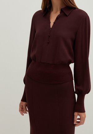 Vínová halenka s výstřihem do V na knoflíky, žebrovaným pasem a dlouhými rukávy s přiléhavými manžetami, vyrobená z hladké tkaniny.