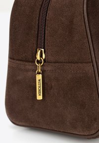 Bruine suède tas met een gouden rits en een merketiket, met een gladde textuur, afgeronde vorm en subtiele stiksels.