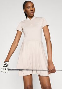 Beiges Golfkleid mit kurzen Ärmeln, das einen Reißverschlusskragen und einen ausgestellten Rock hat. Getragen mit einem weißen Golfhandschuh und einem Golfschläger.