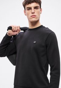 Čierna mikina s okrúhlym výstrihom z bavlny, s malým logom na hrudi a hladkým, textúrovaným povrchom. Model drží tašku.