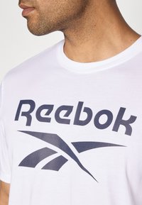 Weißes Baumwoll-T-Shirt mit großem navyblauen "Reebok"-Logo und Grafikdesign, Rundhalsausschnitt und kurzen Ärmeln.
