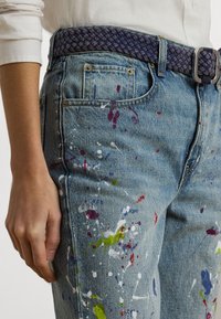 Blue denim jeans with multicolored paint splatters, featuring a braided navy textile belt and silver buckle, shown on a mannequin's side.
