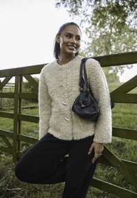 Giacca beige pelosa con tre bottoni frontali, abbinata a una borsa nera. Il modello indossa pantaloni neri larghi, in piedi contro una recinzione di legno.