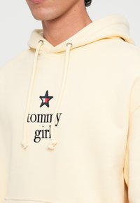 Sudadera de color amarillo claro hecha de material suave, con un parche de estrella negra con un acento rojo y el texto bordado "tommy girl" en la parte frontal.
