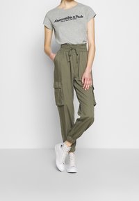Grå bomulds t-shirt med sort skrift, olivenfarvede cargo-bukser med elastiske manchetter og hvide høj-hælede sneakers, stående på en lys baggrund.