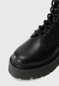 Black leather boot featuring a rounded toe, sturdy sole with deep treads, and black laces. Detailed stitching accents the design.