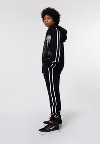 Enfant debout, de profil, portant un hoodie noir et un pantalon noir avec des rayures blanches, ainsi que des baskets montantes noires et blanches.