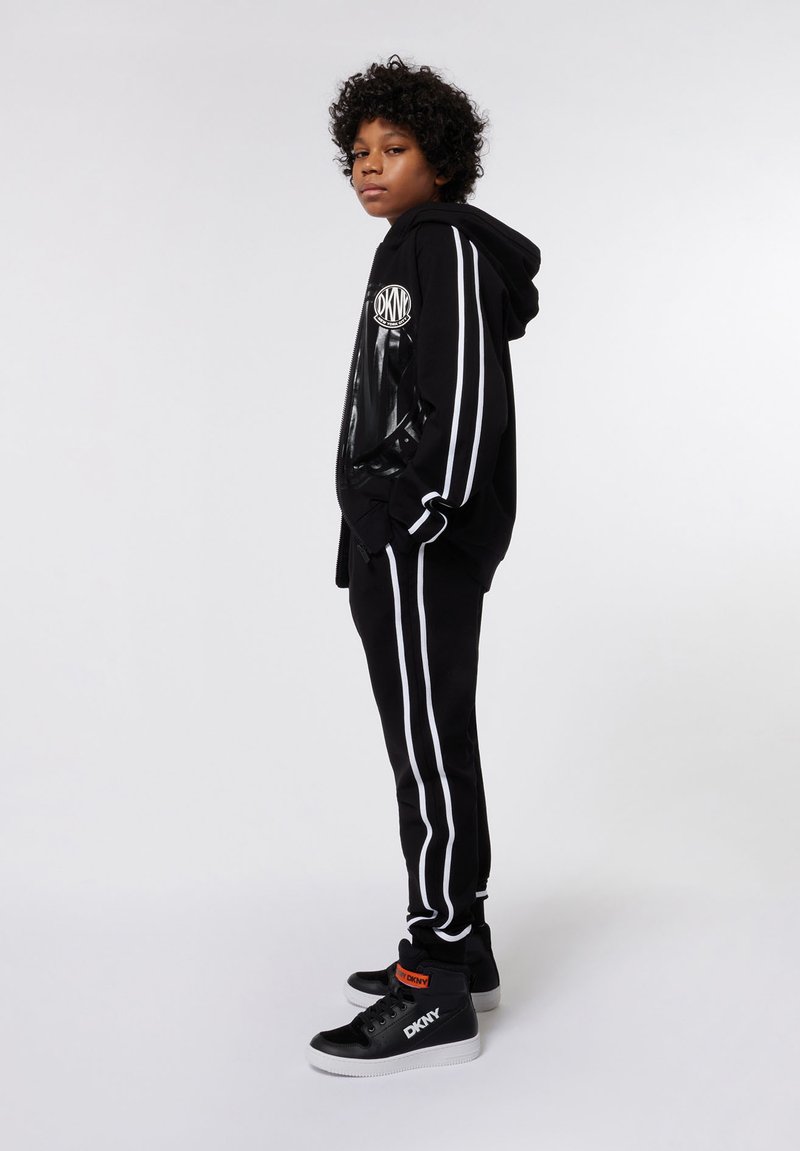 Enfant debout, de profil, portant un hoodie noir et un pantalon noir avec des rayures blanches, ainsi que des baskets montantes noires et blanches.