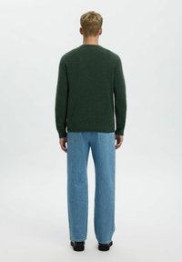 Maglione verde lavorato a maglia con maniche lunghe e scollatura tonda, abbinato a jeans a gamba larga blu chiaro e calzature nere. Vista da dietro.