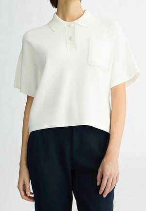 Personne portant un polo en maille blanc à manches courtes avec des boutons et un détail de poche texturé, associé à un pantalon foncé.