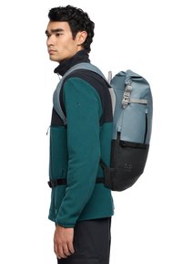 Grau-blauer Rucksack mit verstellbaren Trägern und schwarzem Boden, ausgestattet mit einem Rollverschluss und einer seitlichen Reißverschlusstasche, getragen von einer Person in einer blauen Jacke.