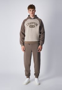 Felpa marrone e beige con grafica "CHAMPION PHYS ED", abbinata a pantaloni della tuta marroni coordinati. Il tessuto sembra morbido; il modello indossa scarpe da ginnastica bianche.