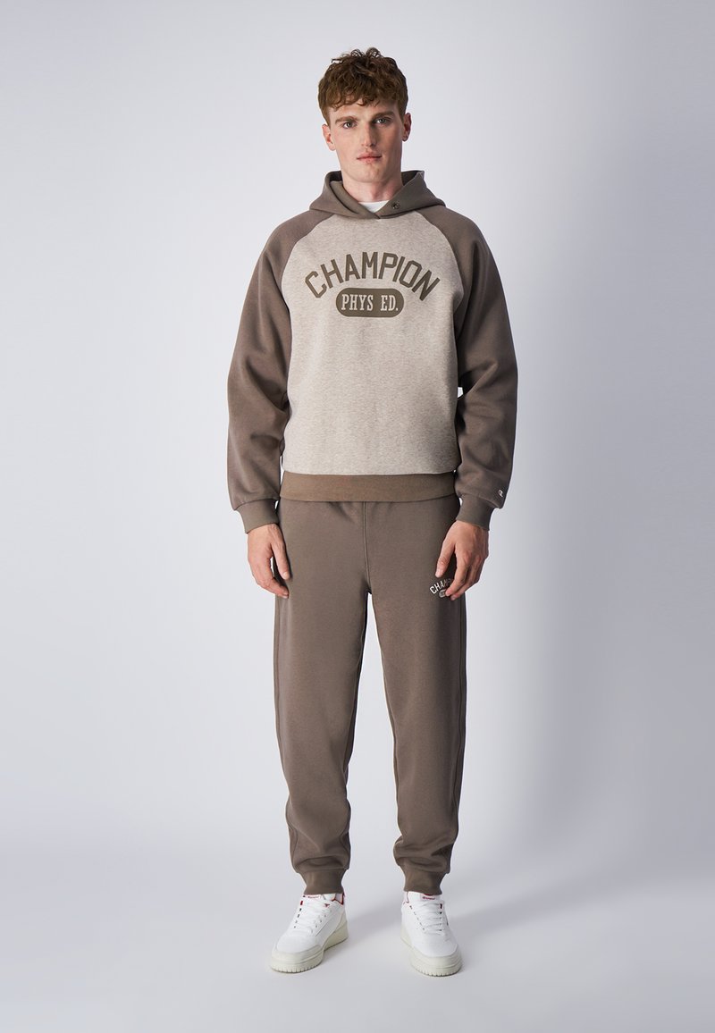 Felpa marrone e beige con grafica "CHAMPION PHYS ED", abbinata a pantaloni della tuta marroni coordinati. Il tessuto sembra morbido; il modello indossa scarpe da ginnastica bianche.