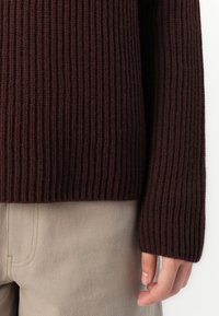 Maglione bordeaux a coste con maniche lunghe, caratterizzato da un design a maglia testurizzato. Il maglione è abbinato a pantaloni beige chiaro.