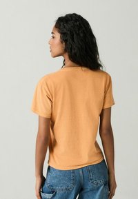 Kurzärmliges T-Shirt in einem sanften Orangeton, mit lockerer Passform und schlichtem Ausschnitt, kombiniert mit hochgeschnittenen Jeans.