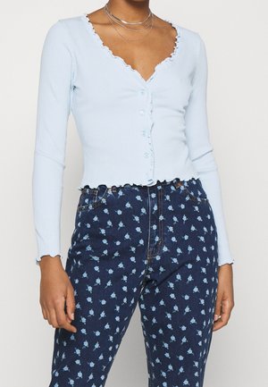 Nainen, jolla on päällään vaaleansininen napillinen neule, jossa on aaltoilevat reunat, ja tummansiniset farkut, joissa on pieniä vaaleansinisiä kukkia.