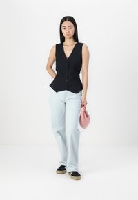 Black sleeveless vest with a v-neck and five buttons, paired with light blue flared jeans and a small pink handbag. Black shoes with a tan sole.