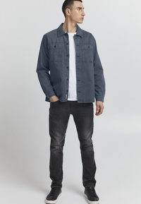 Mørkeblå skjorte med knapper og to frontlommer, kombinert med hvit t-skjorte, svarte jeans og svarte joggesko. Glatt stofftekstur.
