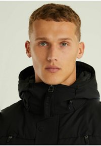 Schwarze Kapuzenjacke mit hohem Kragen, ausgestattet mit Reißverschlusstaschen und einem Logotag auf der Vorderseite. Glatte, matte Textur.