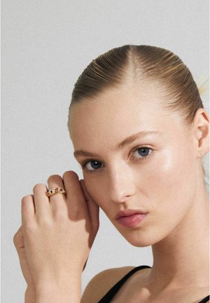 Bague en or avec des maillons entrelacés et un accent en pierre précieuse unique, portée sur une main devant un fond neutre. Texture lisse et polie.