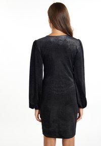 Robe noire longueur genoux avec de longues manches bouffantes et une texture scintillante. Présente un col rond et une silhouette ajustée.