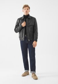 Giacca di pelle nera con zip frontale, due tasche sul petto e una finitura liscia. Indossata con jeans blu scuro e scarpe beige, in piedi su uno sfondo bianco.