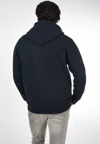Marineblauwe hoodie met een grote, verstelbare capuchon, ribgebreide manchetten en zoom, gemaakt van zachte stof. De achterkant toont een naadloos ontwerp en eenvoudige vorm.