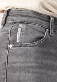Sive traperice s visokim strukom, suptilnom teksturom, bočnim džepovima i etiketom s natpisom "Marc O'Polo Denim" na prednjem džepu.