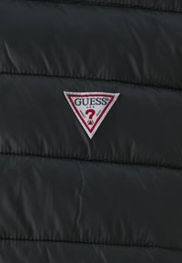 Juodas pūkinis švarkas su siuvinėtu tekstūra. Yra trikampio formos emblema balta ir raudona su "GUESS" prekės ženklu.