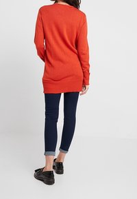 Oranje trui met lange mouwen en geribbelde zoom, gedragen met donkere blauwe skinny jeans en zwarte schoenen, gezien vanaf de achterkant.