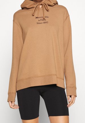 Beżowa bluza z kapturem ze ściągaczami i napisem „Abercrombie & Fitch Since 1892”, noszona z czarnymi przylegającymi szortami rowerowymi.