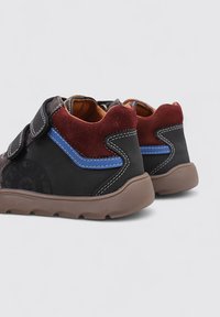 Zapatos de cuero negro y ante con una suela marrón, detalles en azul y rojo, y tiras de Velcro. Presentan materiales texturizados y detalles cosidos.